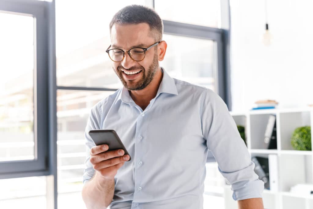 Image closeup of smiling employer guy in white shirt standing in office room near big window and using smartphone for work